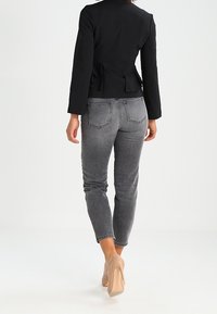 Černá bunda s peplum detailem, která je kombinována se světle šedými úzkými džíny. Modelka má na sobě nahé lodičky. Plné pozadí, zaměření na oblečení.