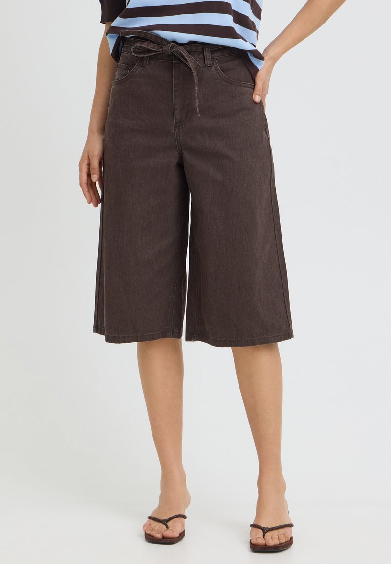 Femme portant un short large brun foncé longueur genou avec une ceinture nouée, associé à un haut rayé et des tongs noires.