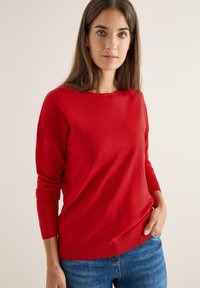 Rode gebreide trui met lange mouwen en een ronde halslijn. Gladde textuur en geribde manchetten en zoom. Gepaard met blauwe jeans.