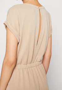 Beige jumpsuit met een keyhole-detail aan de achterkant, korte opgerolde mouwen en een elastische taille met een geplooide textuur. Gladde stofafwerking.