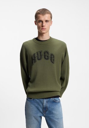Ung mand iført en olivengrøn ribbet sweater med teksten "HUGO" og lyseblå jeans, stående mod en ensfarvet hvid baggrund.