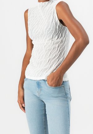 Mouwloze witte top met textuur, gecombineerd met lichtblauwe spijkerbroeken, ene hand in de zak, op een persoon met een huidtint van gemiddelde kleur tegen een witte achtergrond.