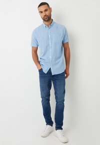Camicia a maniche corte azzurro chiaro con bottoni e taschino sul petto, abbinata a jeans blu scuro e sneakers bianche, in piedi su uno sfondo neutro.