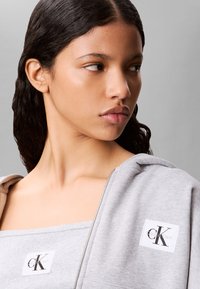 Joven con cabello oscuro que lleva una sudadera con cremallera gris de Calvin Klein y un top tubo a juego, mirando hacia su derecha sobre un fondo liso.