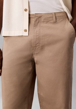 Nærbilde av person som har på seg beige bukser og en hvit strukturert skjorte med knapper, med en hånd i lommen.