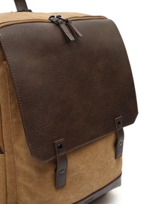 Mochila de lona con una solapa de imitación cuero marrón, acentos oscuros y dos botones laterales; presenta un acabado texturizado y detalles cosidos.