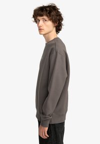 Sudadera de algodón gris oscuro con puños y dobladillo de canalé, cuello redondo y un sutil detalle de logo en el frontal, combinada con jeans oscuros.