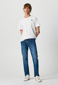Camiseta blanca de algodón con mangas cortas, que presenta un pequeño logo negro en el pecho. Combinada con unos vaqueros de denim azul y zapatillas blancas.