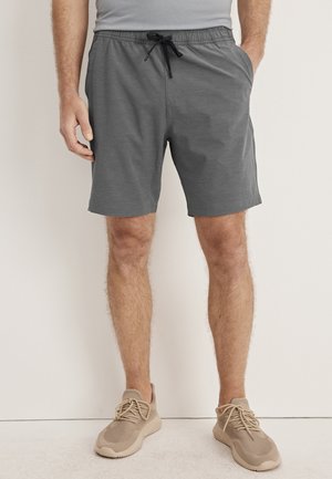 Man wearing gray drawstring shorts and beige knit sneakers standing against a white wall with one hand in pocket.