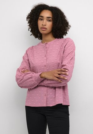 Blouse rose clair texturée avec des manches bouffantes, un col rond, des boutons de fermeture et des embellissements floraux sur le tissu.