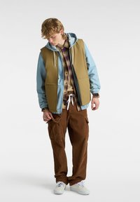 Bruine corduroy broek, gelaagd met een beige vest over een blauwe jas en een geruit overhemd. Witte sneakers en een trekkoordaccent op de broek.