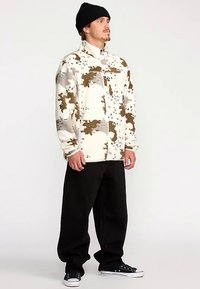 Witte fleecejas met bruine en grijze abstracte patronen, volledig met rits sluiting, hoge kraag, gecombineerd met zwarte losse broek en zwarte sneakers.