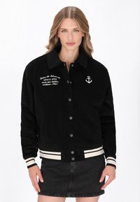 Czarna kurtka z korduroju z kołnierzem, z przodu na guziki, białym haftem kotwicy i paskowymi mankietami. Tekst na klatce piersiowej dodaje szczegółu.