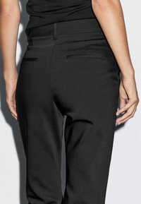 Schwarze maßgeschneiderte Hose mit glatter Textur, die einen hohen Bund und zwei Gesäßtaschen aufweist. Keine sichtbaren Verschlüsse oder Verzierungen.