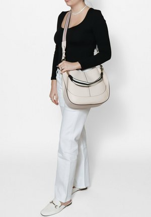 Beige leren handtas met een zwart-witte band, gebogen vorm en contrasterende stiksels. Model draagt een zwarte top en witte broek.