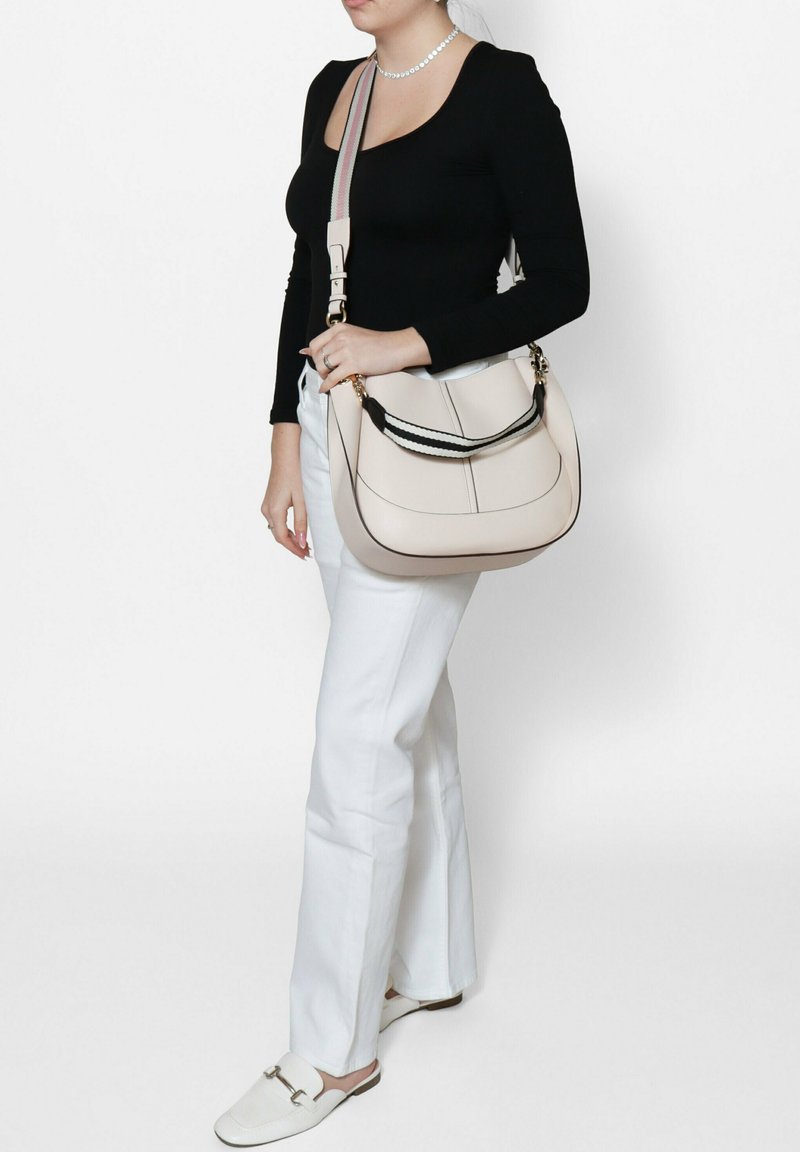 Borsa in pelle beige con tracolla bianca e nera, forma curva e dettagli di cucitura a contrasto. Il modello indossa una cima nera e pantaloni bianchi.