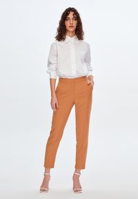 Weißes Hemd mit Kragen, taillierte Passform, kombiniert mit orangen Slim-Fit-Hosen. Sandalen mit Knöchelriemen runden den Look ab. Einfache helle Hintergrund.