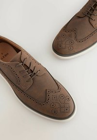 Zapatos brogue marrones hechos de cuero liso y malla, con perforaciones decorativas, punta redonda y suela de goma blanca.