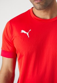Camiseta deportiva roja hecha de tejido suave, con escote redondo y mangas cortas. Logo blanco de Puma en el pecho izquierdo, detalle de malla en la manga.