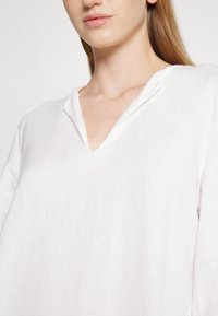 Femme portant un haut ample en lin blanc avec un col en encoche, laissant voir son cou et une partie de son visage et de son épaule.