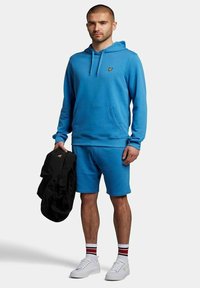 Felpa di cotone blu con tasca frontale, abbinata a pantaloni corti coordinati. L'outfit presenta un piccolo logo giallo e sneakers bianche con calzini a strisce rosse e blu.