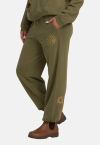 Oliwkowe dresy w luźnym kroju, z złotymi motywami słońca na lewym udzie i dolnej części nogawki. Wykonane z miękkiej, teksturowanej tkaniny.