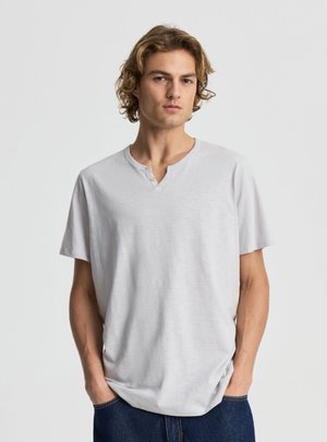 Giovane uomo con capelli biondi ricci indossa una maglietta henley grigio chiaro a maniche corte e jeans blu scuro, in piedi con le mani in tasca.