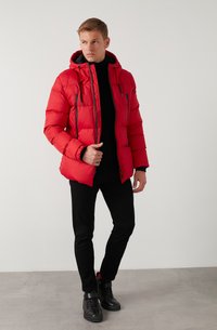 Rød polstret jakke med hætte, sorte lynlåsdetaljer og elastiske manchetter. Båret over en sort turtleneck, parret med sort slim-fit bukser og støvler.