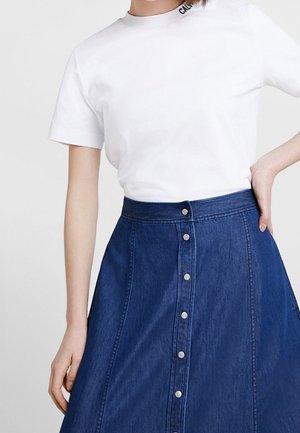 Mujer con camisa blanca de manga corta y falda vaquera azul de talle alto con botones plateados en la parte delantera.