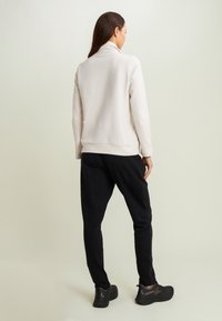 Donna in piedi di spalle che indossa una maglietta bianca a maniche lunghe, pantaloni neri e sneakers nere su uno sfondo chiaro e uniforme.