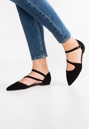 Pies con jeans ajustados azules que llevan zapatos planos negros puntiagudos con doble correa, de pie sobre un fondo blanco.