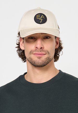Giovane uomo con barba chiara che indossa un berretto beige con logo EA7 nero e oro e una maglietta verde scuro con scollo a girocollo, sfondo neutro.