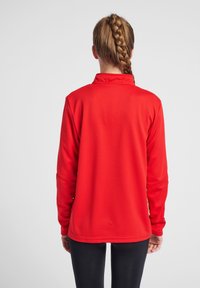 Maglietta sportiva rossa a maniche lunghe con finish testurizzato, colletto e design aderente, vista dalla schiena. Abbinata a leggings neri.