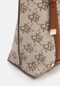 Nærbillede af hjørnet af en beige totebag med brunt DKNY-logo mønster, brune stropper og metaldetaljer i guldfarve på hvid baggrund.