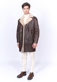 Abrigo de cuero marrón con un cuello de borrego, con cierre de botones, bolsillos laterales y una textura suave. Lleva puesto un cuello alto color crema.