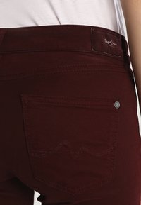Burgundy jeans med en bakficka som har en vågigt sydd design och en liten metallknapp, tillverkade av ett mjukt material.