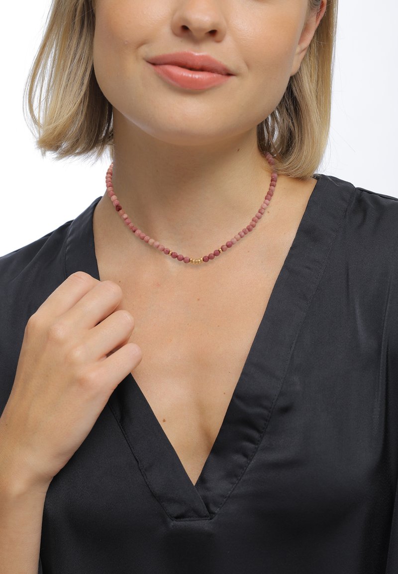 Perlekæde med pink og lilla sten samt guldfarvede detaljer, sat mod en sort v-hals top. Glat tekstur og rund form.