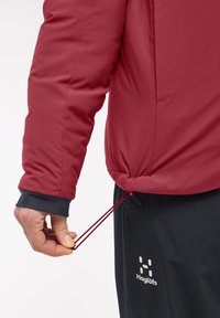 Veste isolante rouge avec des poignets élastiques, dotée d'un cordon de serrage à l'ourlet. Associée à un pantalon noir arborant un logo de marque blanc.