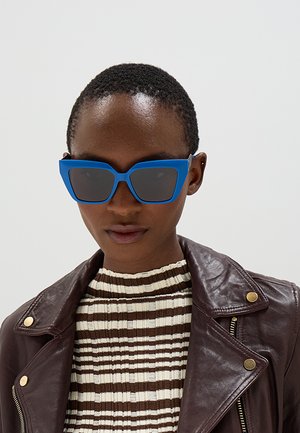 Persona con capelli corti che indossa grandi occhiali da sole blu, top a righe marrone e bianco e giacca di pelle marrone scuro con bottoni dorati.