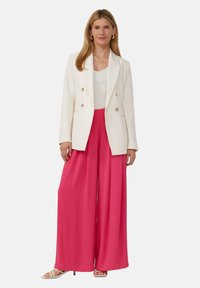 Weiße doppelte Blazerjacke über einem taillierten weißen Oberteil, kombiniert mit lockeren, leuchtend pinken weit geschnittenen Hosen. Flache weiße Sandalen runden den Look ab.