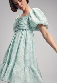Vaaleansininen mekko puffihiuletsilla, rypytetty vyötärö ja levenevä kerroksellinen helma. Teksturoitu kangas esittää kukkakuviota.