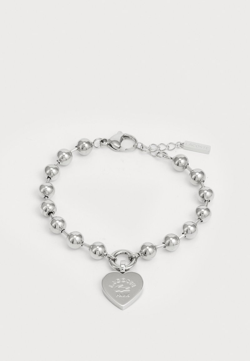 Hõbedane käevõru, millel on ümmargused helmed ja südamekujuline ripats, millel on graveeritud "Lacoste Paris". Kaasas on liblikluku sulgemine.