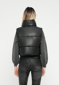 Melnas ādas pūkainā veste ar augstu apkakli, ar gludu tekstūru un quilted dizainu. Valkāta virs pelēka, teksturēta džempera un melnām biksēm.