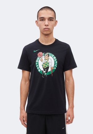 Νέος άνδρας που φοράει μαύρο μπλουζάκι Boston Celtics με λευκορήκονα που περιστρέφει μια μπάλα μπάσκετ, στέκεται μπροστά σε απλό ανοιχτό φόντο.