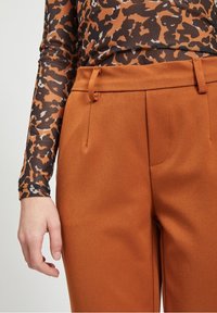Persona vistiendo pantalones de color óxido y una blusa de manga larga con un patrón abstracto en marrón y negro, con la mano relajada a un lado.