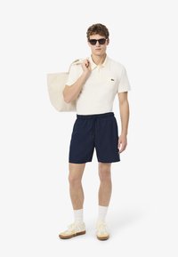 Polo beige con tasca, pantaloni corti blu marino e sneakers beige. La persona porta una borsa tote leggera e indossa occhiali da sole scuri.