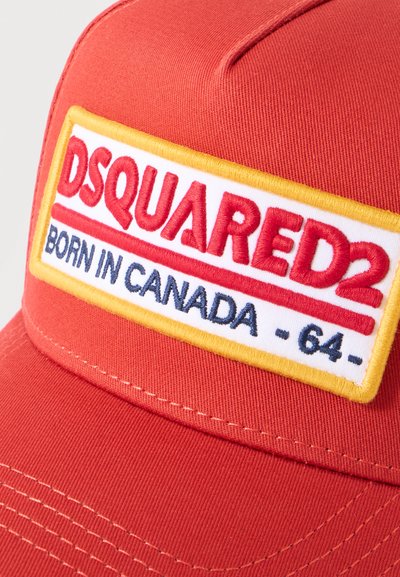 Κόκκινο καπέλο με ορθογώνιο έμβλημα σε κίτρινο περίγραμμα, που εμφανίζει έντονο κόκκινο "DSQUARED2" και μπλε κείμενο "BORN IN CANADA - 64-" κάτω από αυτό.