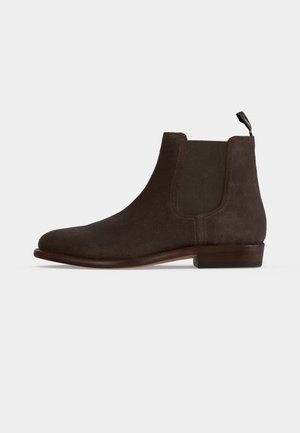Bottine Chelsea en suède marron foncé avec panneaux latéraux élastiques, petit talon empilé et languette à l'arrière sur un fond clair uni.