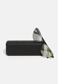Black aviator sunglasses with reflective green lenses, resting on a textured black case featuring embossed branding.