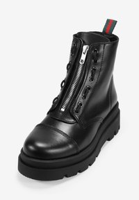 Botín de cuero negro hasta el tobillo con punta redonda, cremallera lateral, cordones decorativos y suela de goma texturizada. Presenta una franja de acento en verde y rojo.